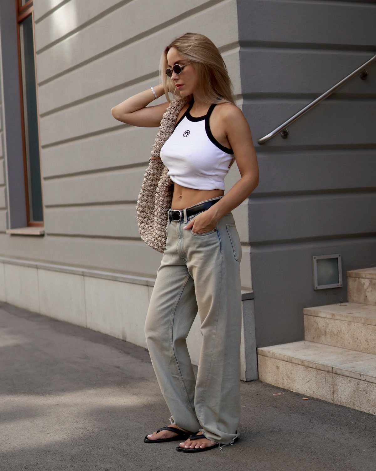 Woman wearing white cropped tank, grey wide-leg trousers, and black flip-flops outdoors.