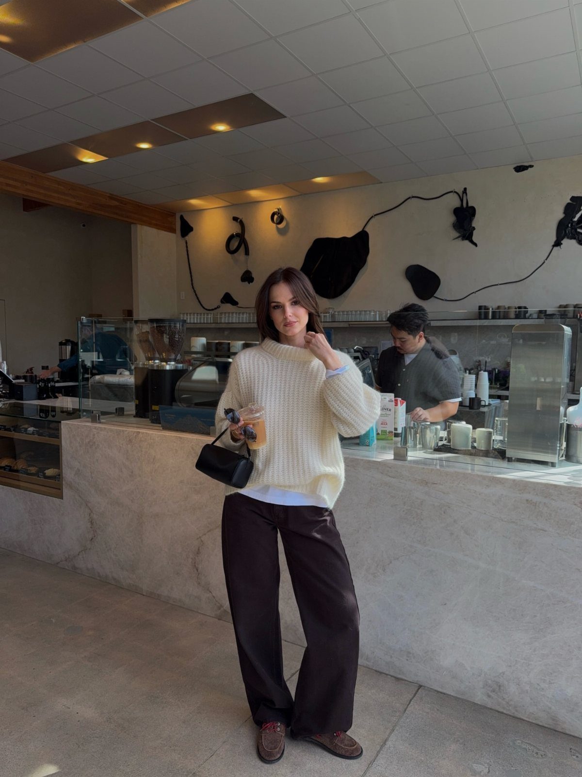 Woman in white textured sweater and brown wide-leg trousers posing in a cafe.