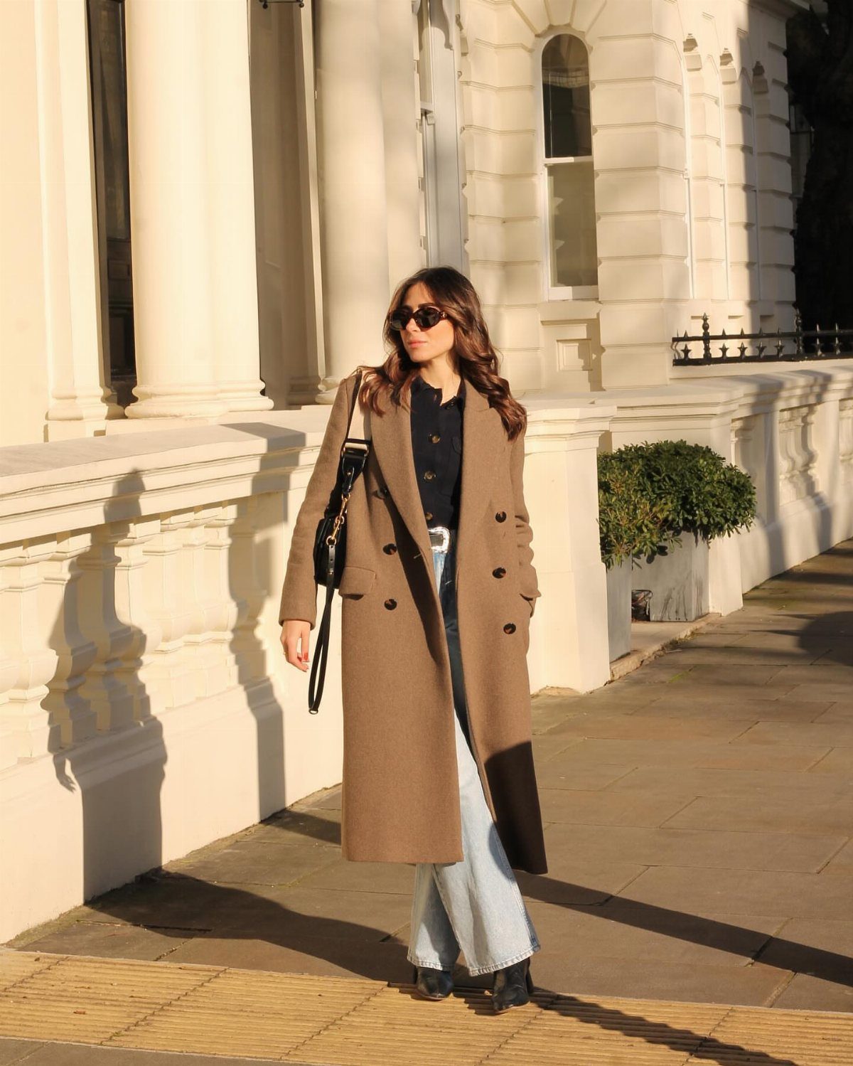 Woman wearing a long tan wool coat over a navy shirt and light jeans.