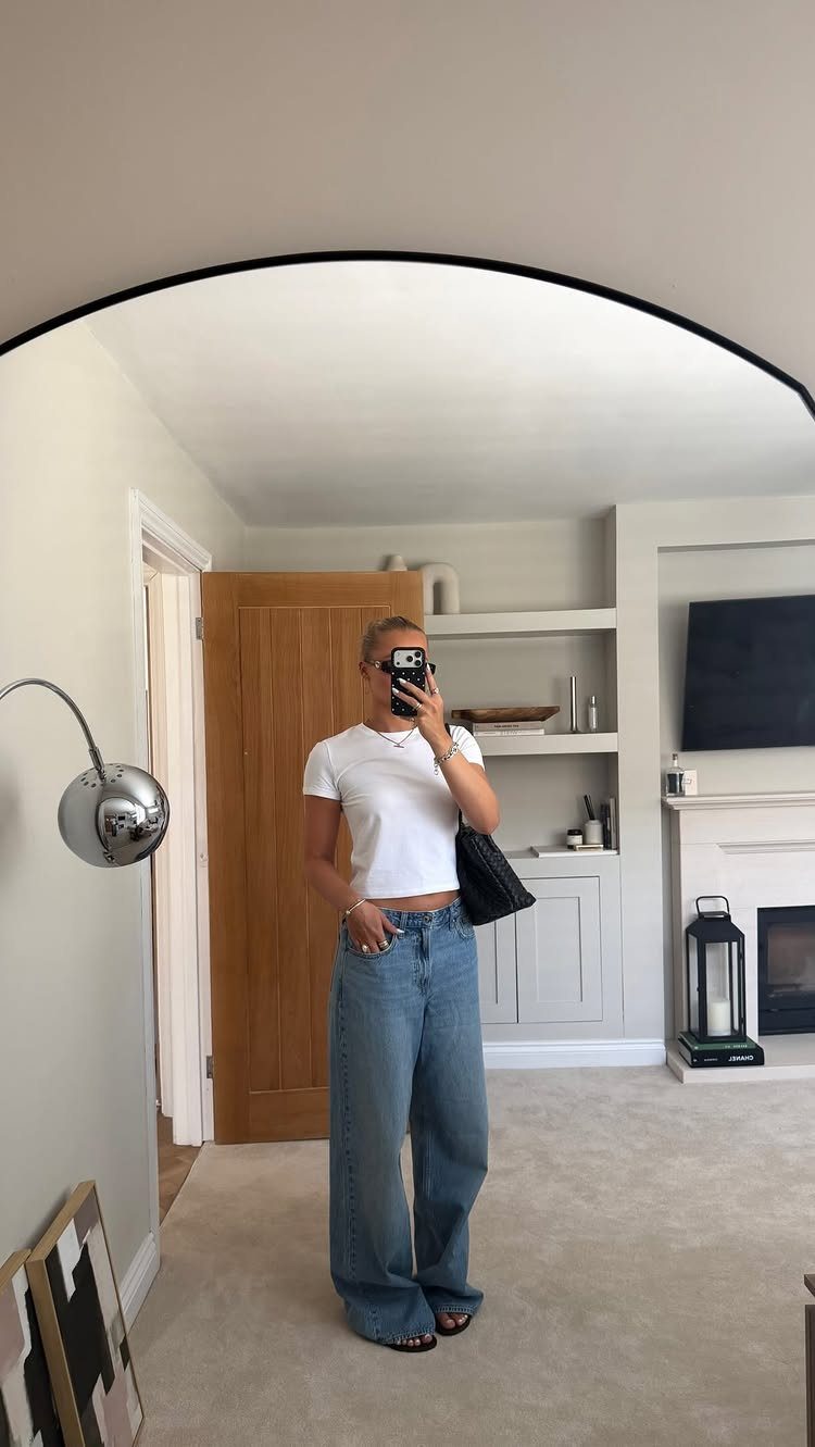 Woman in white t-shirt and wide-leg blue jeans posing for a mirror selfie.
