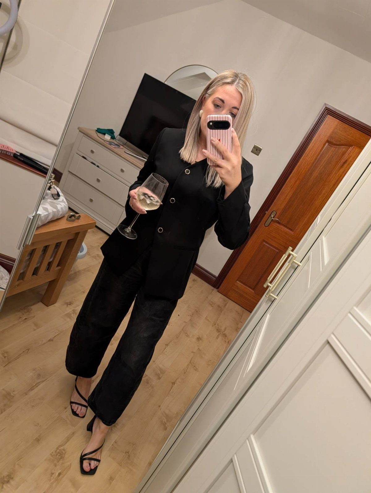 Woman in a black blazer and trousers holding a glass of white wine.