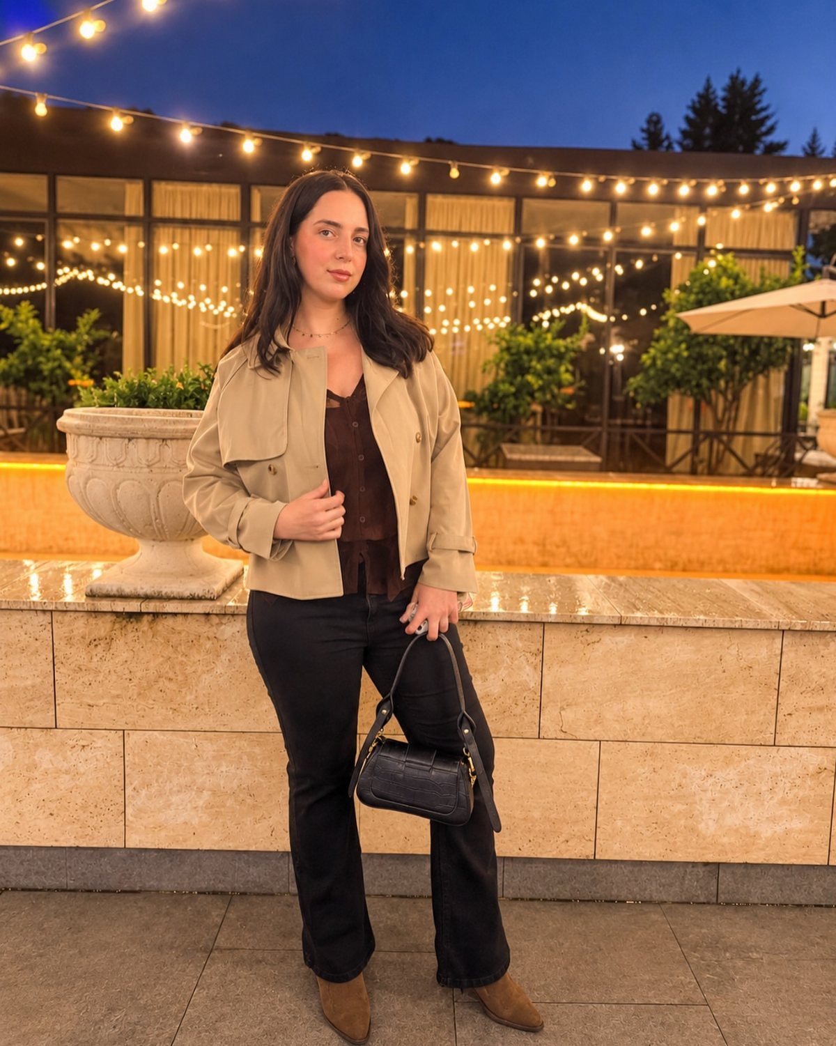 Woman wearing a tan jacket, brown top, and black flared trousers at night.