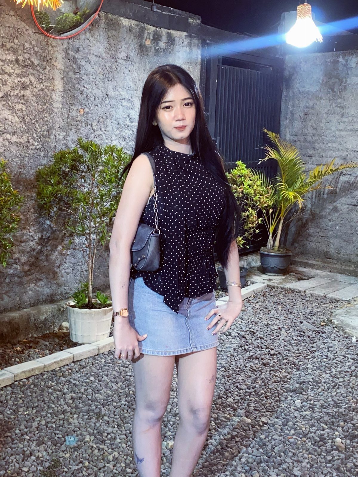 Woman wearing a black polka-dot top and denim mini skirt standing outdoors at night.