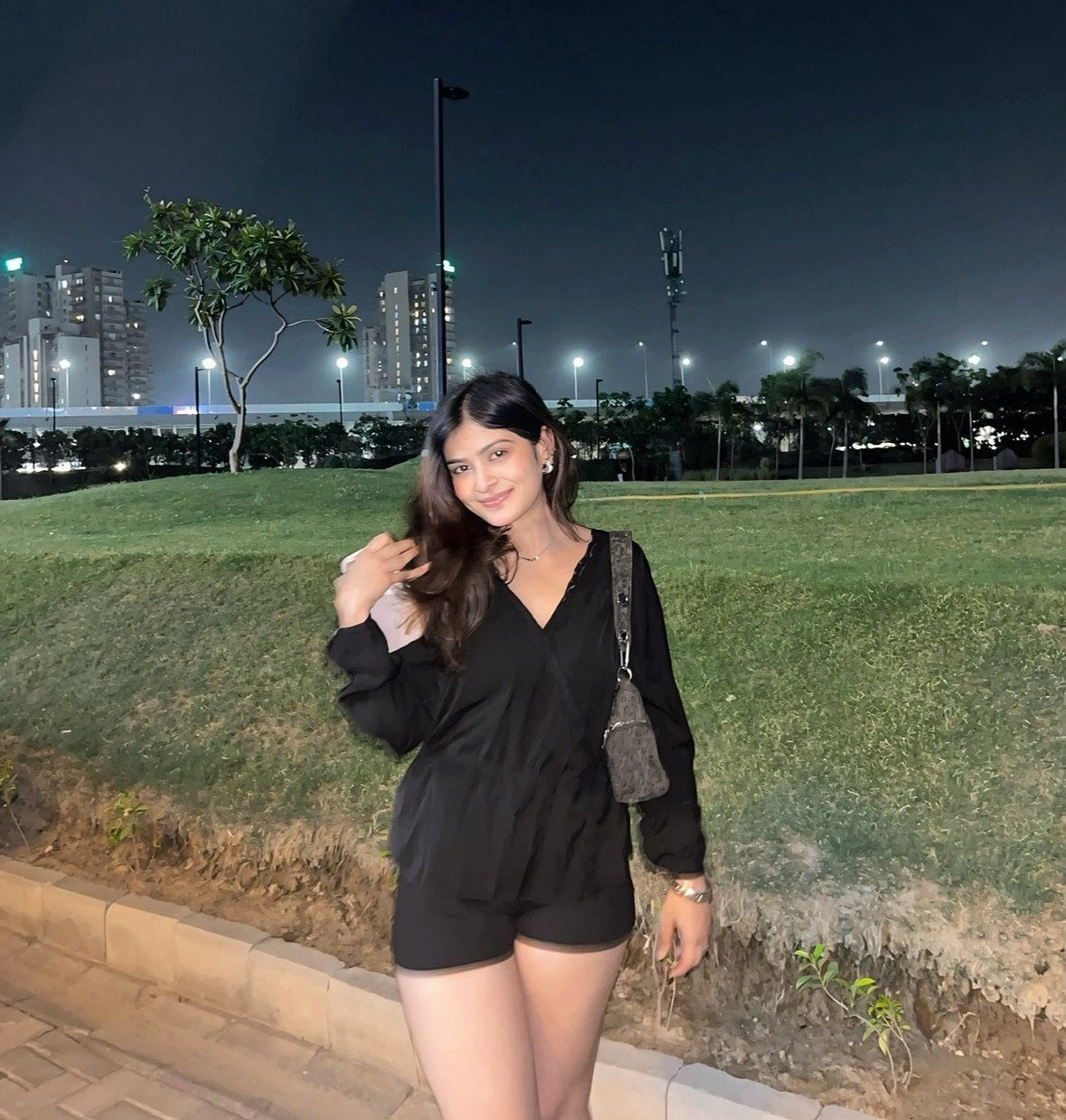 Woman in a black long-sleeved romper and brown bag standing in a park at night.
