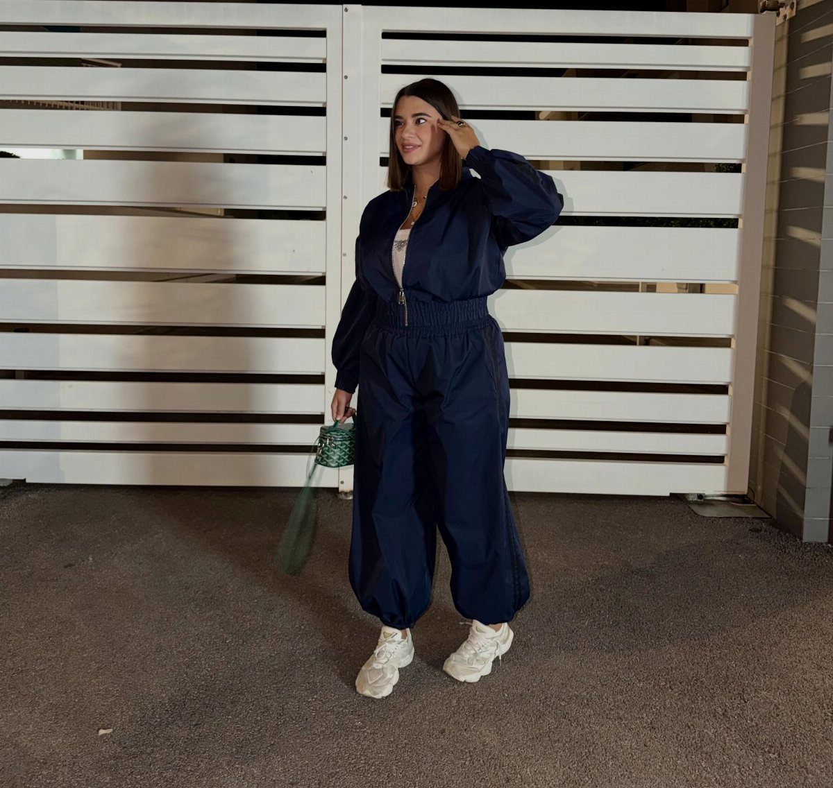 Woman in navy blue jumpsuit and white sneakers standing by a white fence.