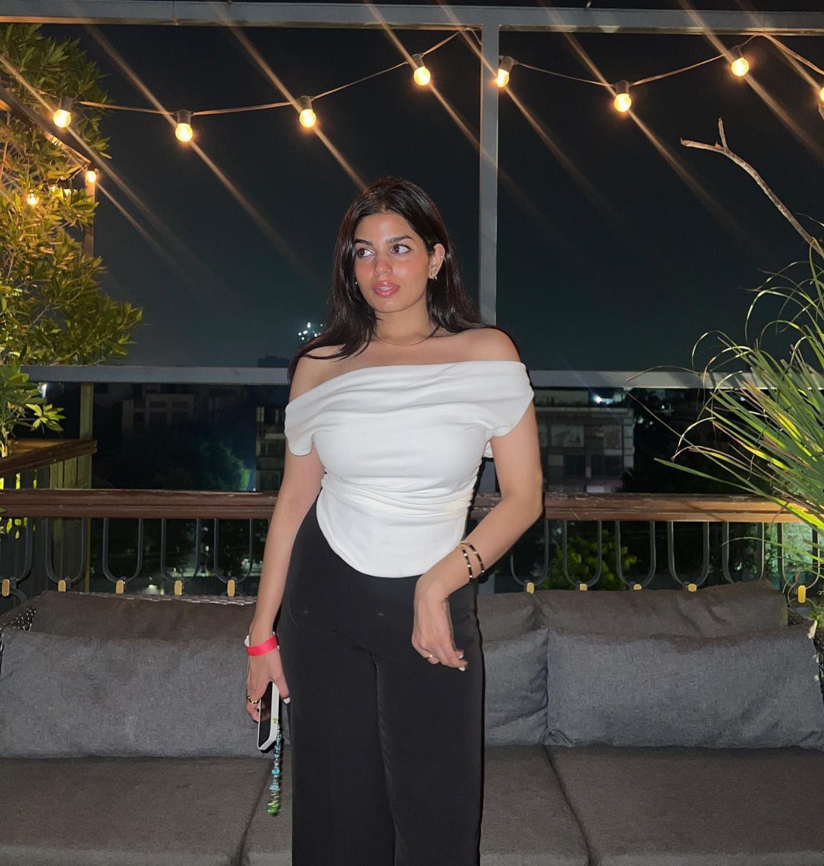Woman in white off-the-shoulder top and black trousers posing on a lit balcony