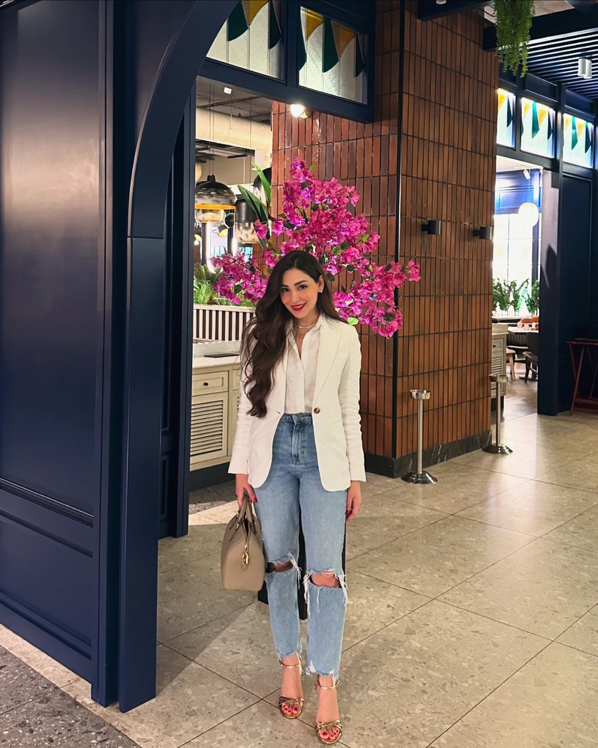 Woman wearing a white blazer and ripped blue jeans with gold strappy heels.