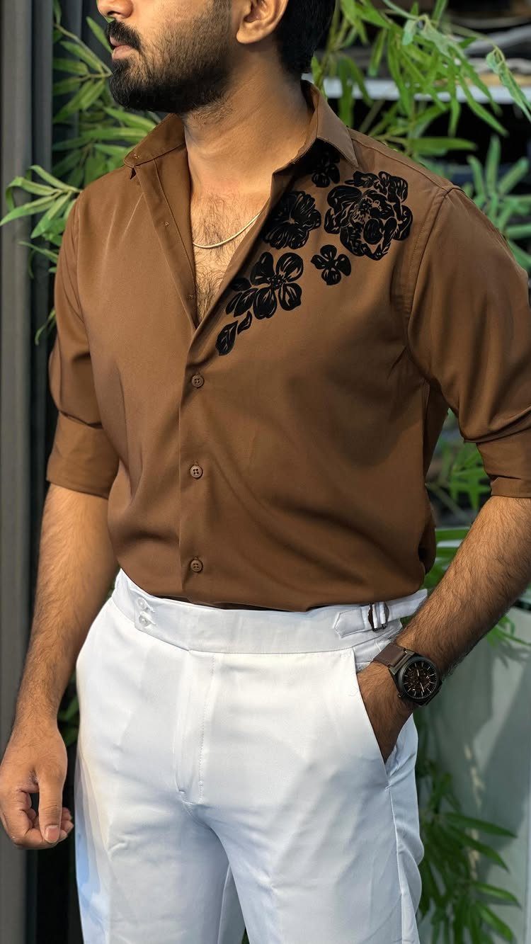 Man in brown embroidered shirt and white trousers posing outdoors with green leaves.