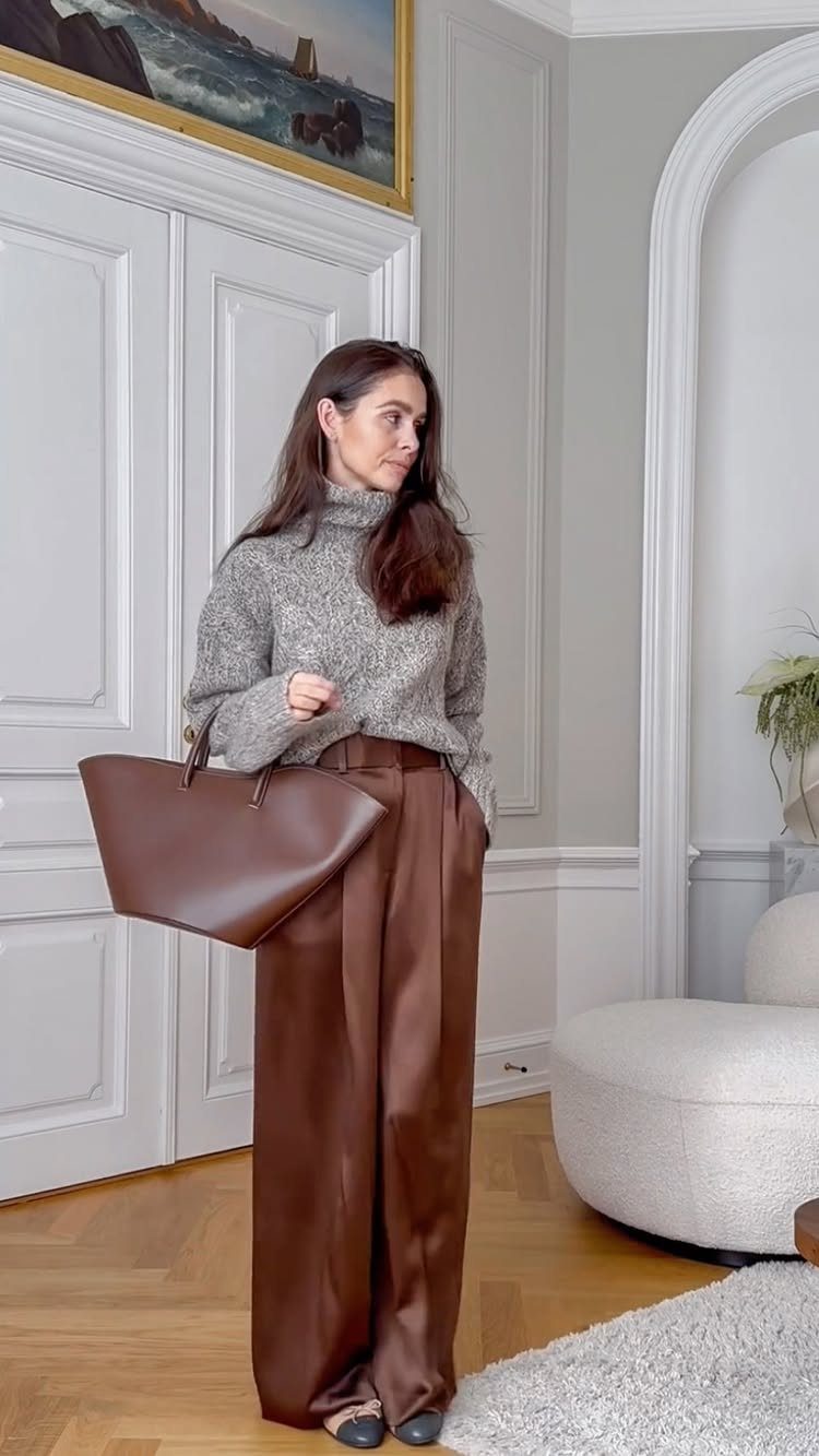 Woman wearing a grey turtleneck sweater and wide-legged brown satin pants with a tote.