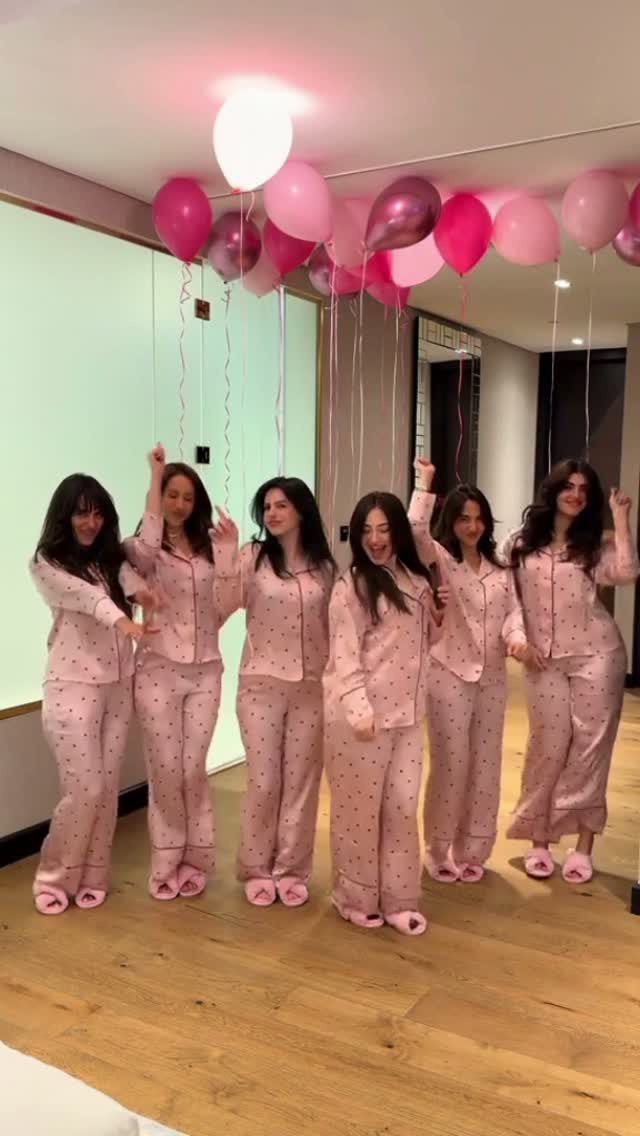 Six women in matching pink pajamas posing under pink and white balloons.