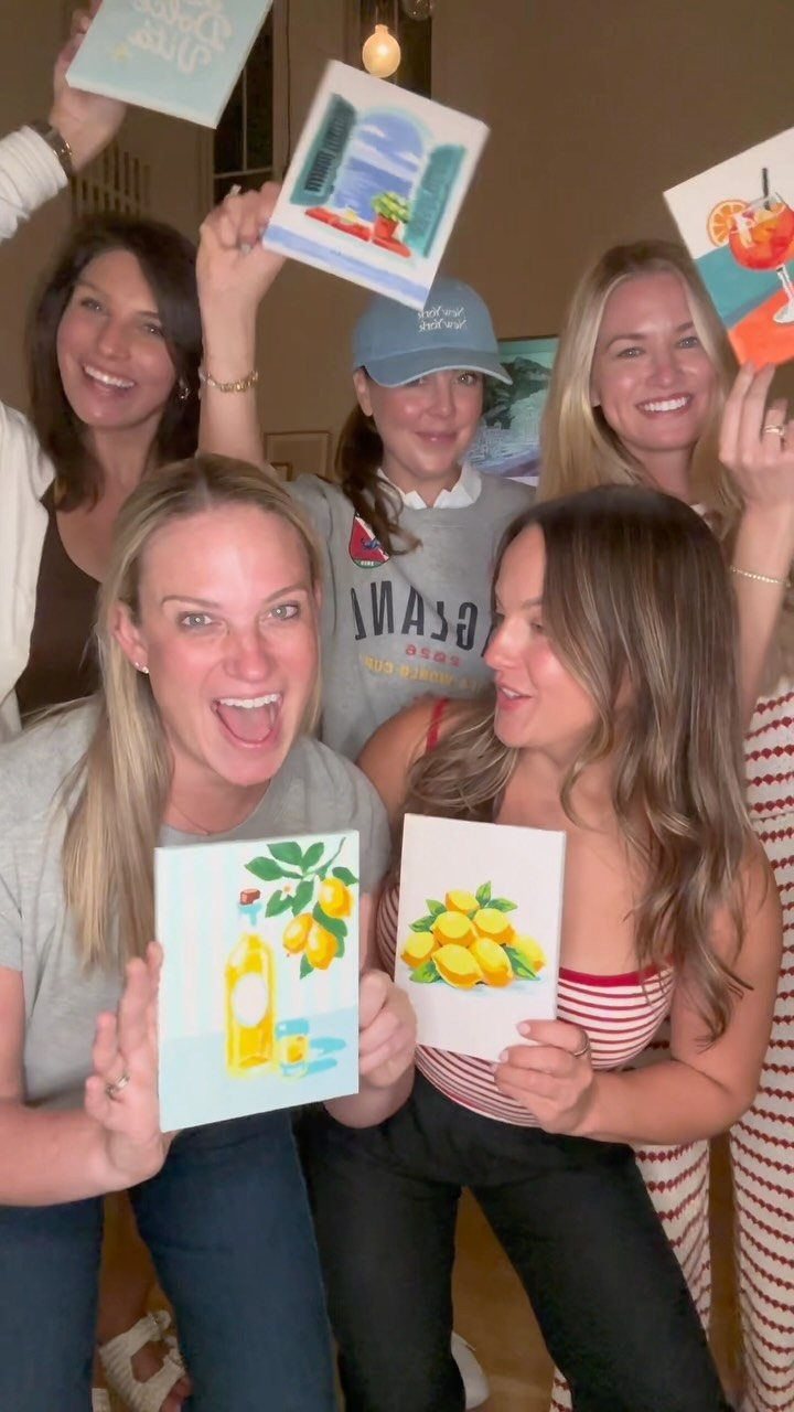 Five women smiling and holding up their small colorful paintings at a party.