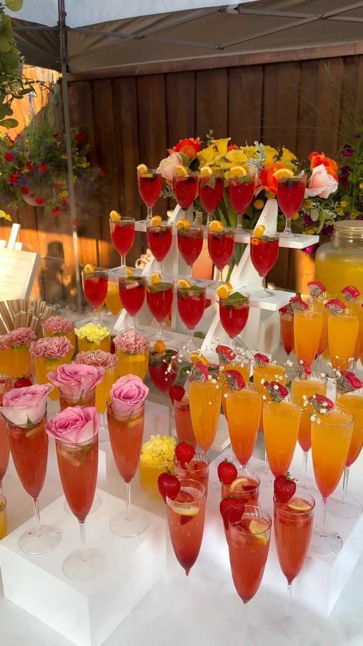 Colorful cocktails with floral garnishes arranged on white tiered stands at an outdoor party.