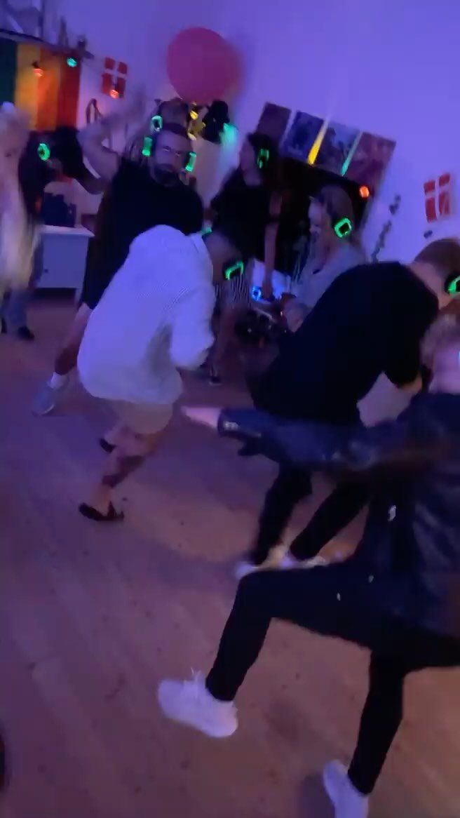 Adults dancing in a purple-lit room wearing glowing green wireless headphones.