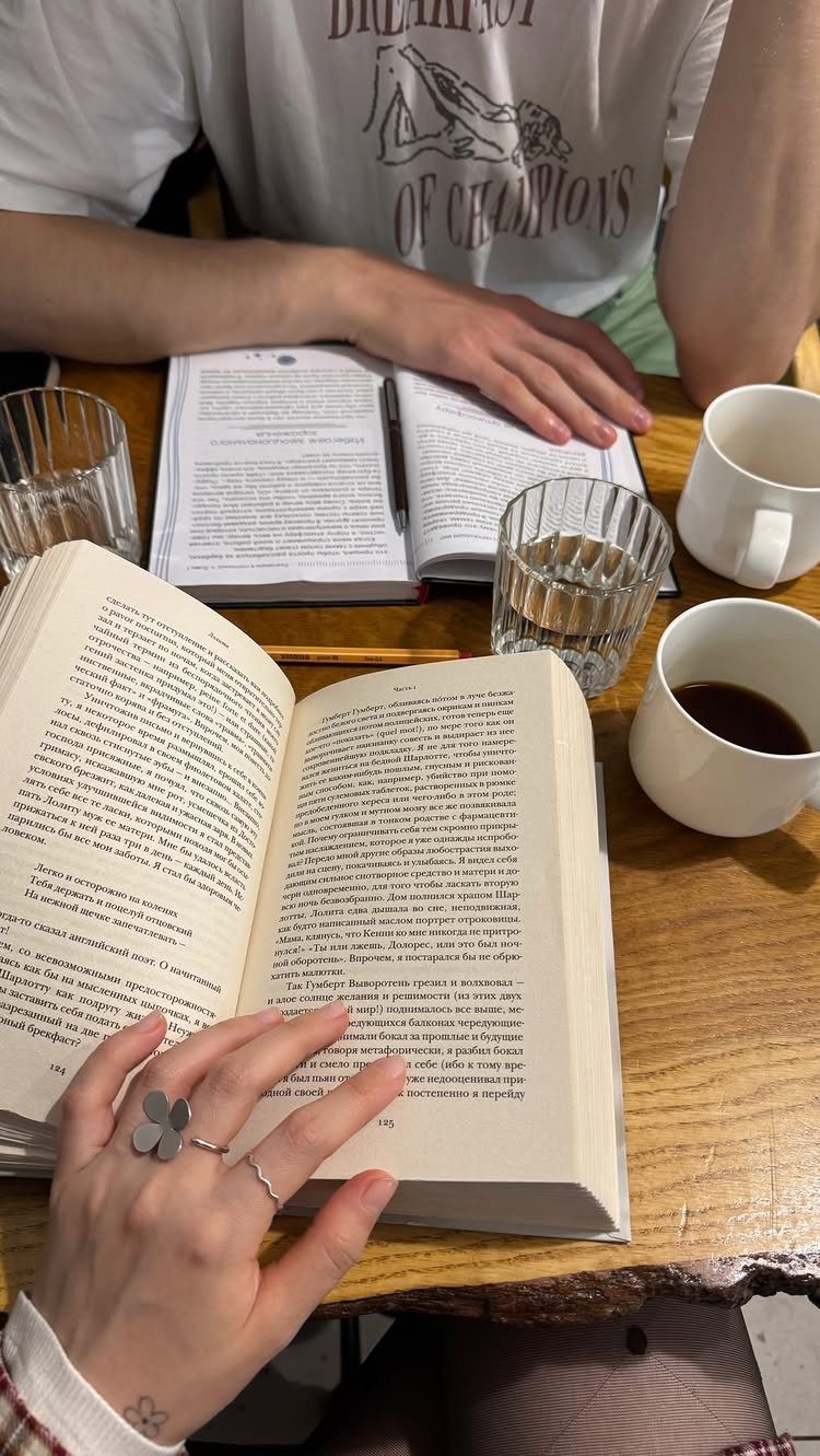 Two people reading books together at a wooden table with coffee cups.