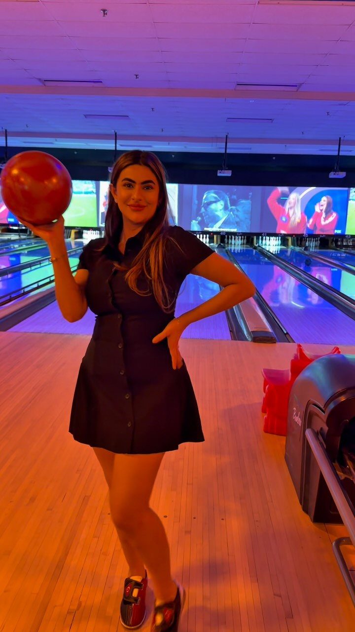 Woman in a black dress holding a red bowling ball at a bowling alley.