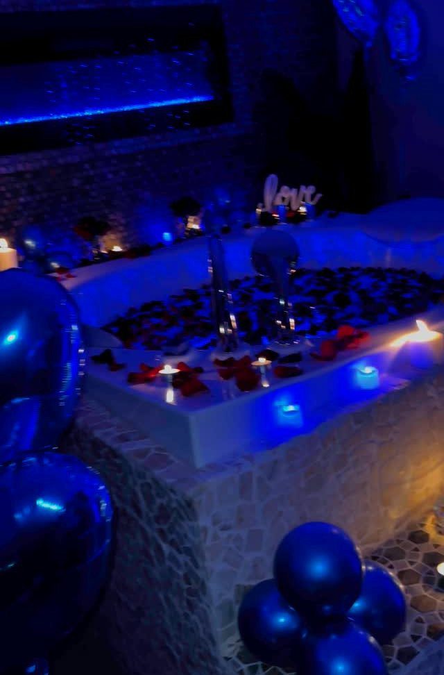 White bathtub filled with red rose petals, blue balloons, and glowing candles.