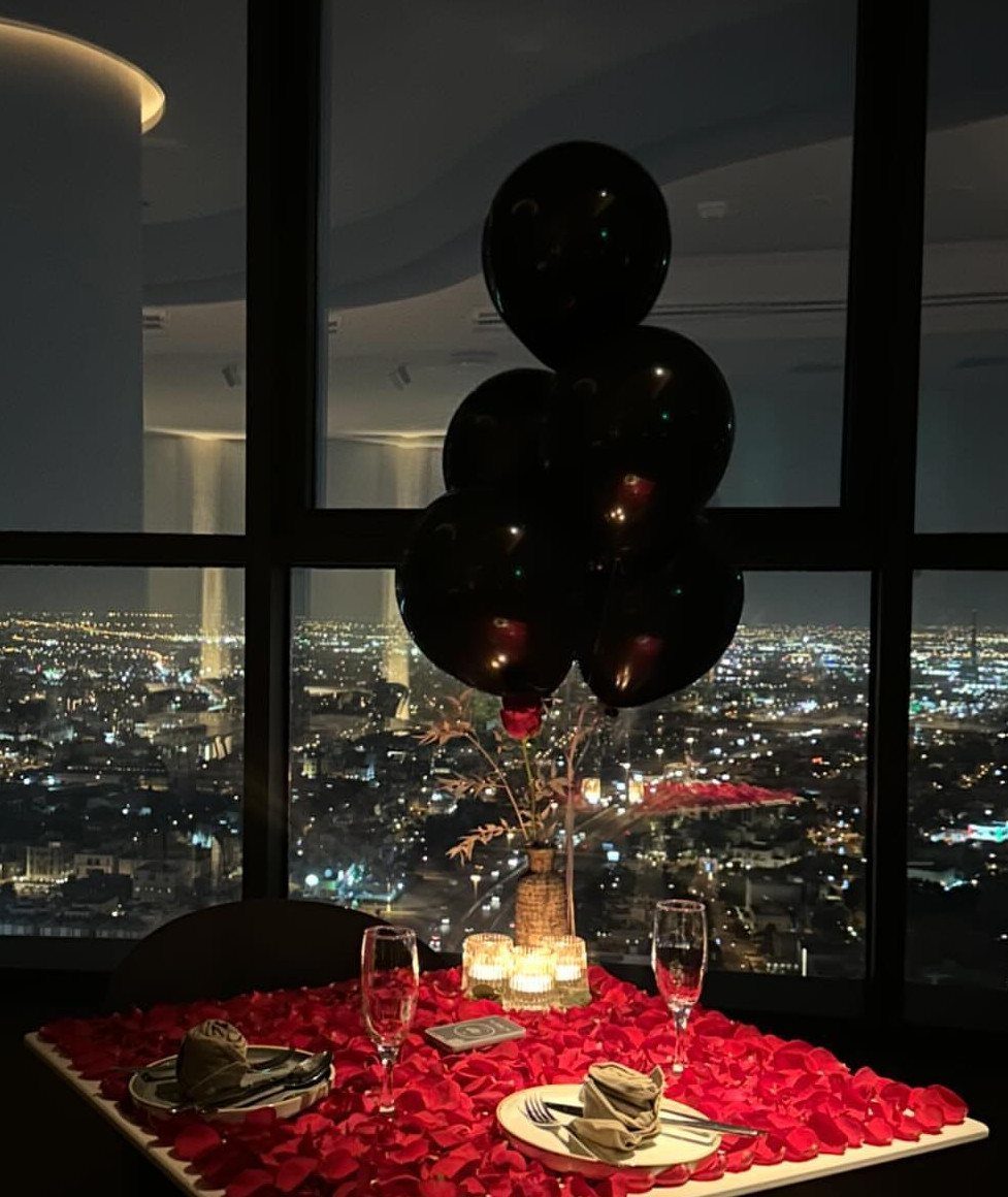 Romantic dinner table with red rose petals and black balloons overlooking a city skyline.
