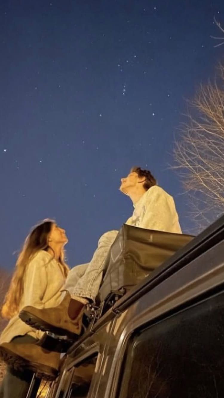 Couple sitting on a car roof looking up at a starry night sky.