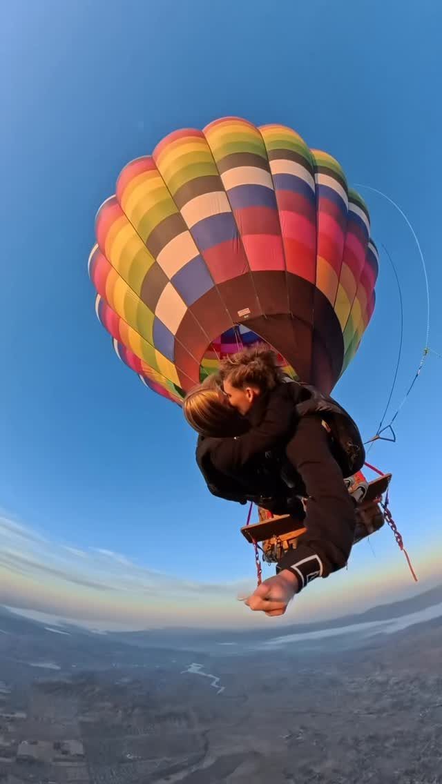 Couple kissing in a rainbow hot air balloon basket over a scenic valley landscape