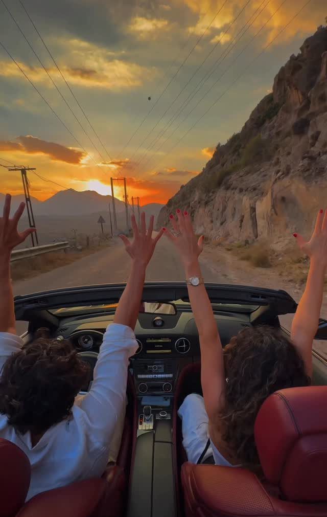 Couple in a red convertible car with arms raised during a mountain sunset drive.