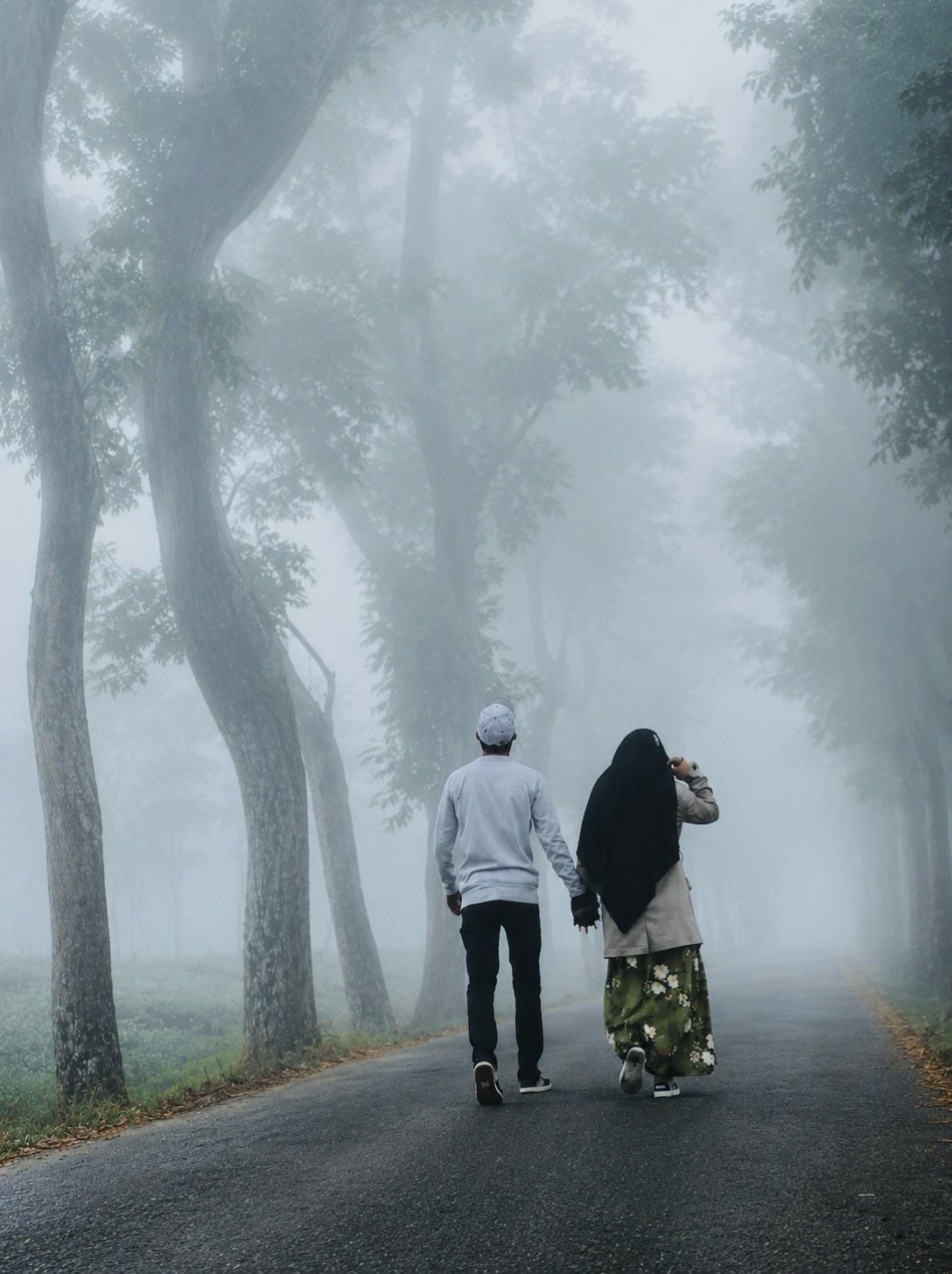 Couple walking hand-in-hand down a foggy road lined with tall trees
