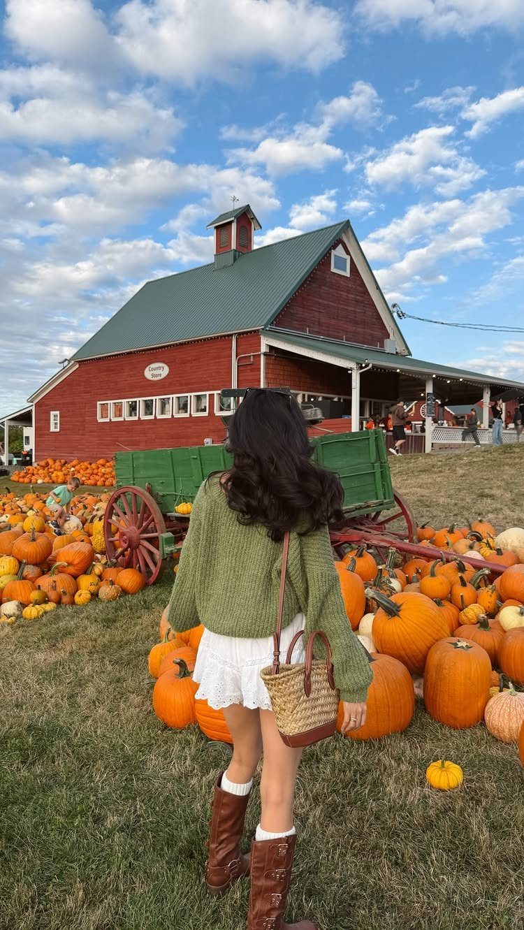 Woman in a green sweater standing in a pumpkin patch before a red barn.