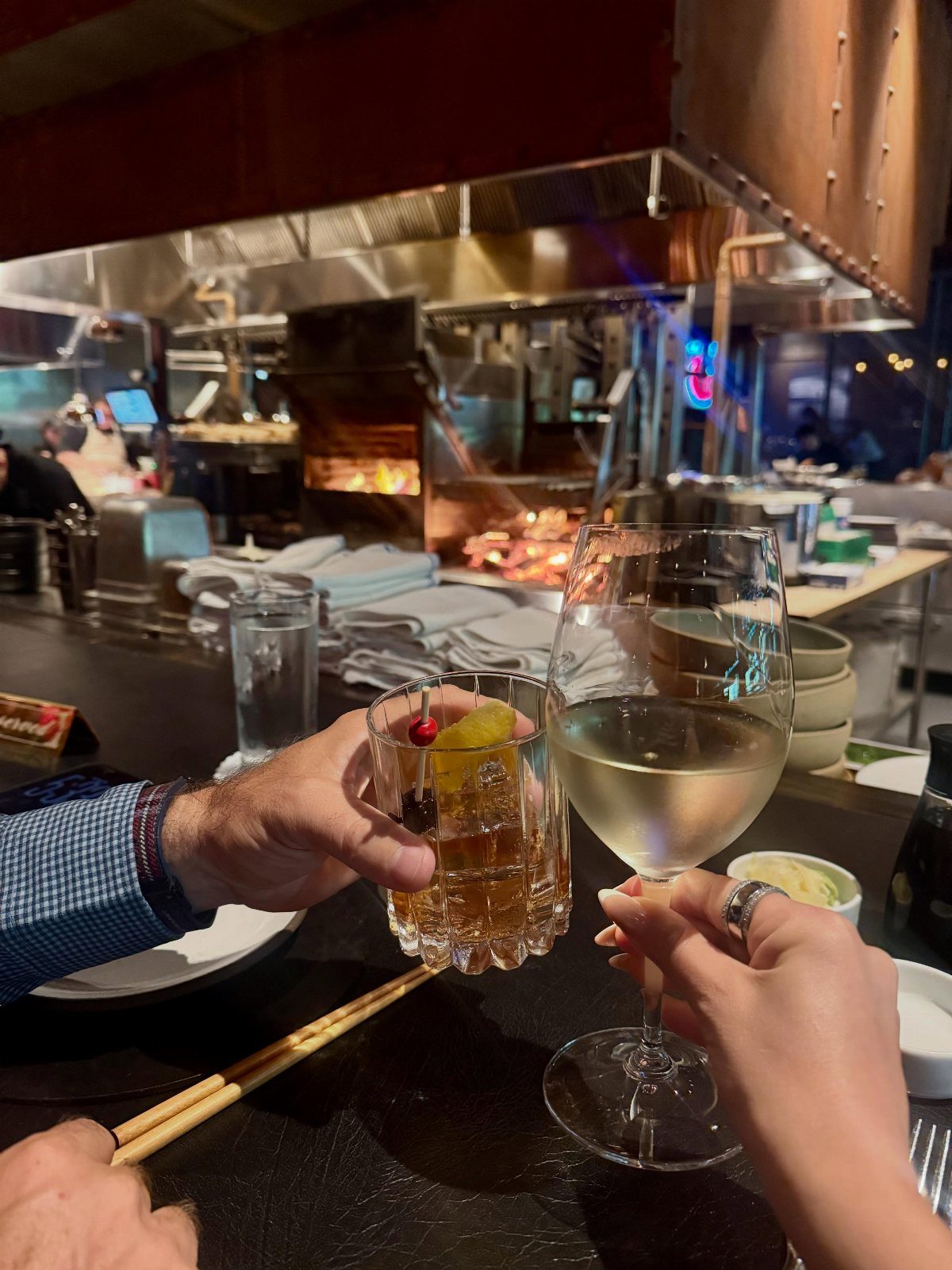 Two people clinking wine and cocktail glasses at a restaurant with open kitchen.
