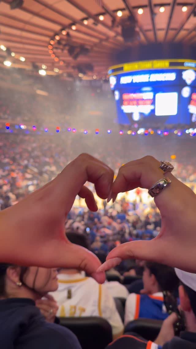 Hands forming a heart shape in front of a crowded sports arena crowd.
