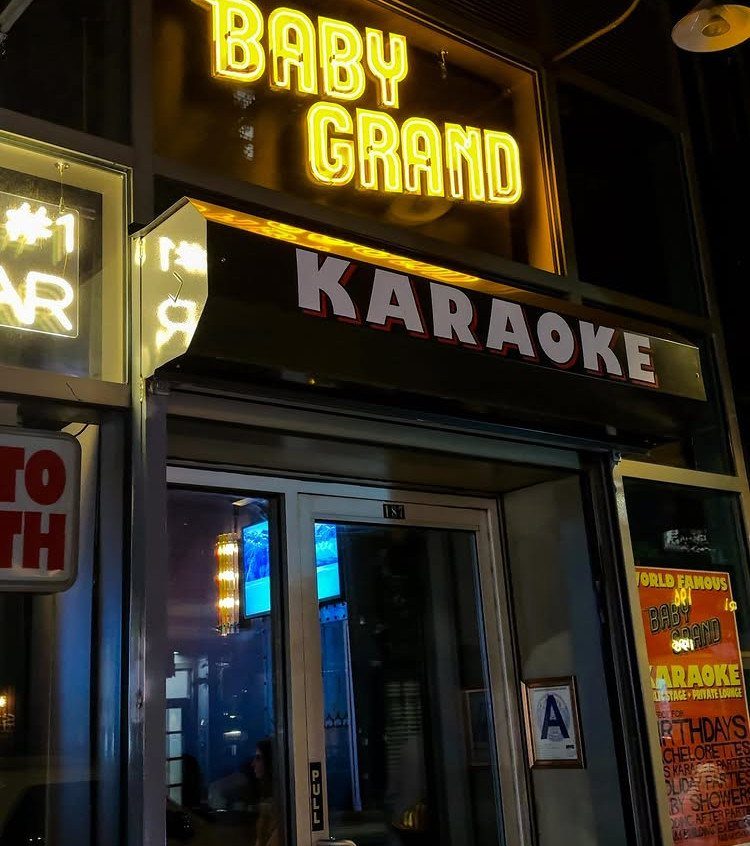 Karaoke bar storefront with yellow neon Baby Grand sign and black awning.
