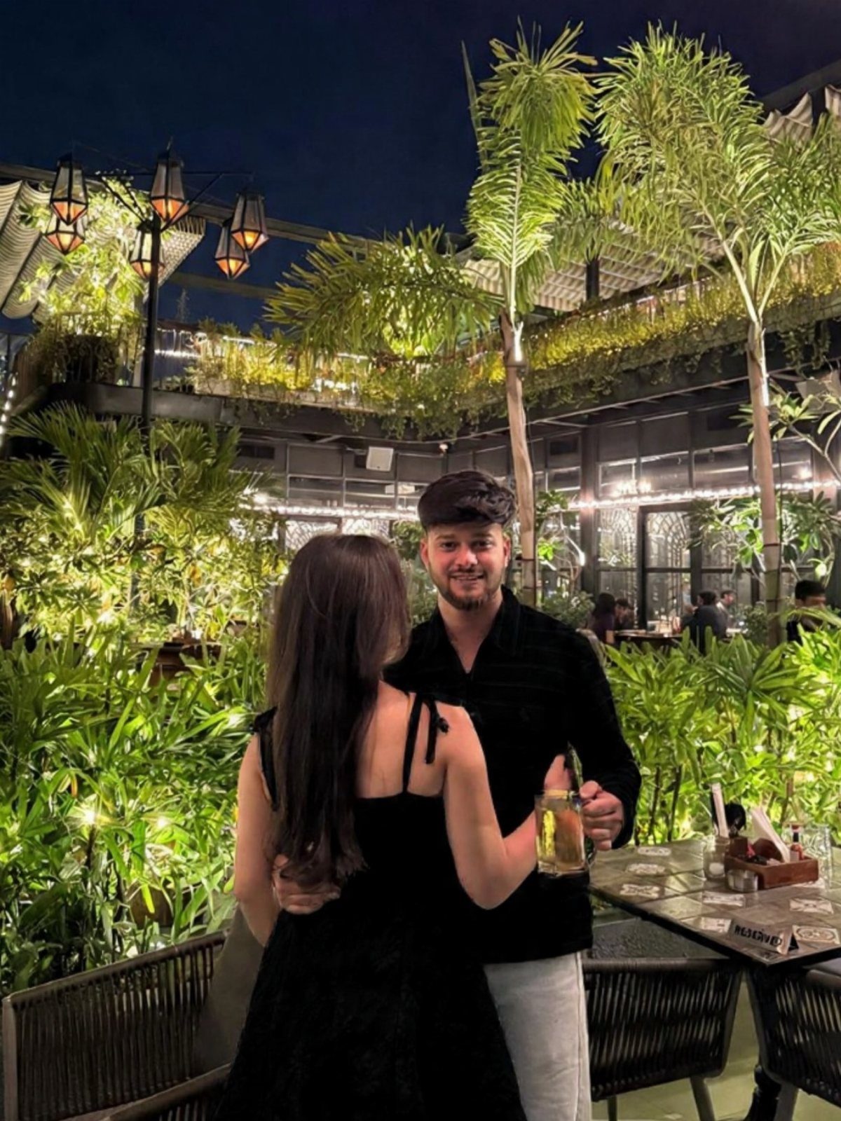 Couple standing together at a lush rooftop bar with drinks and lanterns at night.