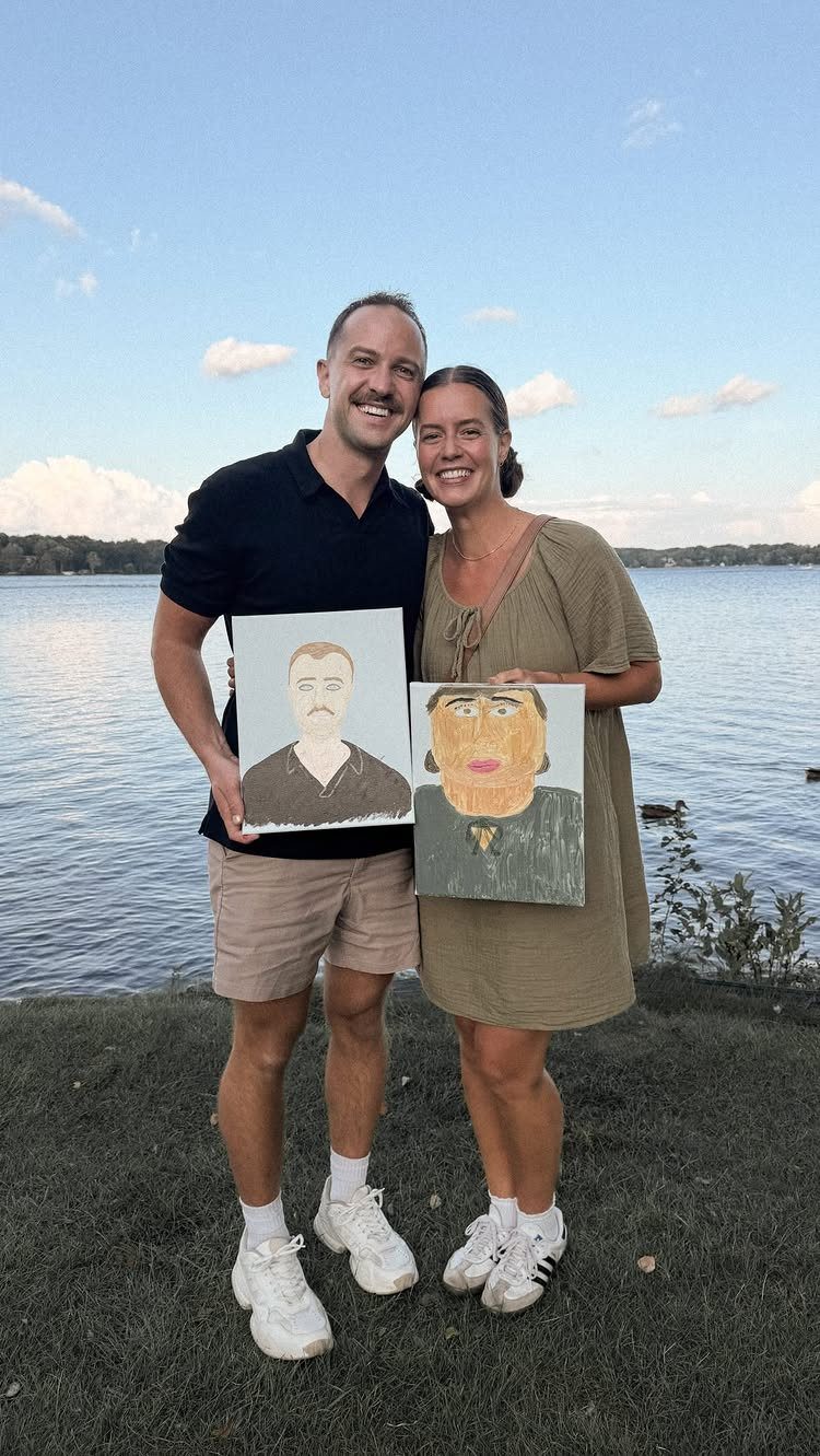 Couple holding hand-painted portraits of each other while standing by a calm lake.