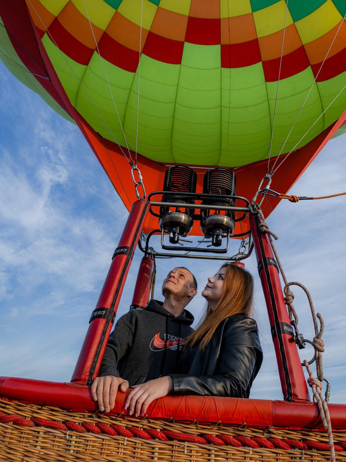 Couple in a wicker basket looking up at a colorful hot air balloon.