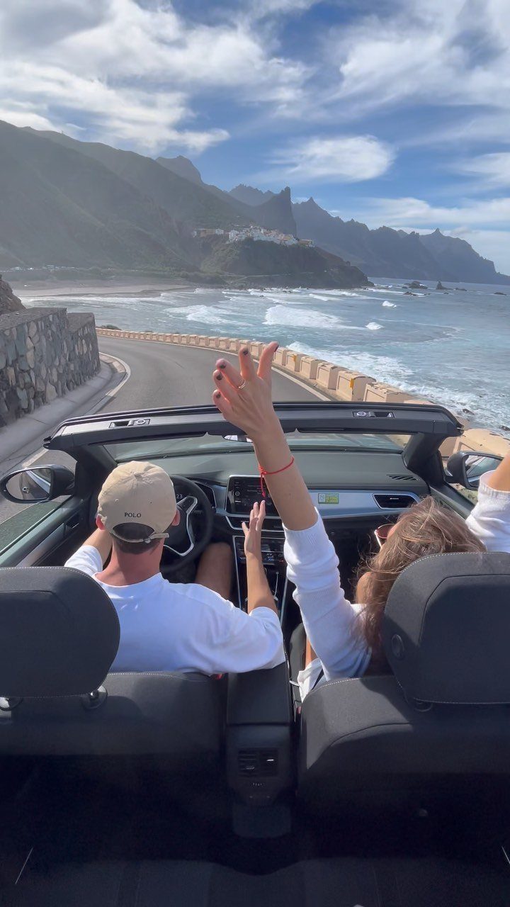 Couple in a white convertible driving along a scenic coastal road with mountains.