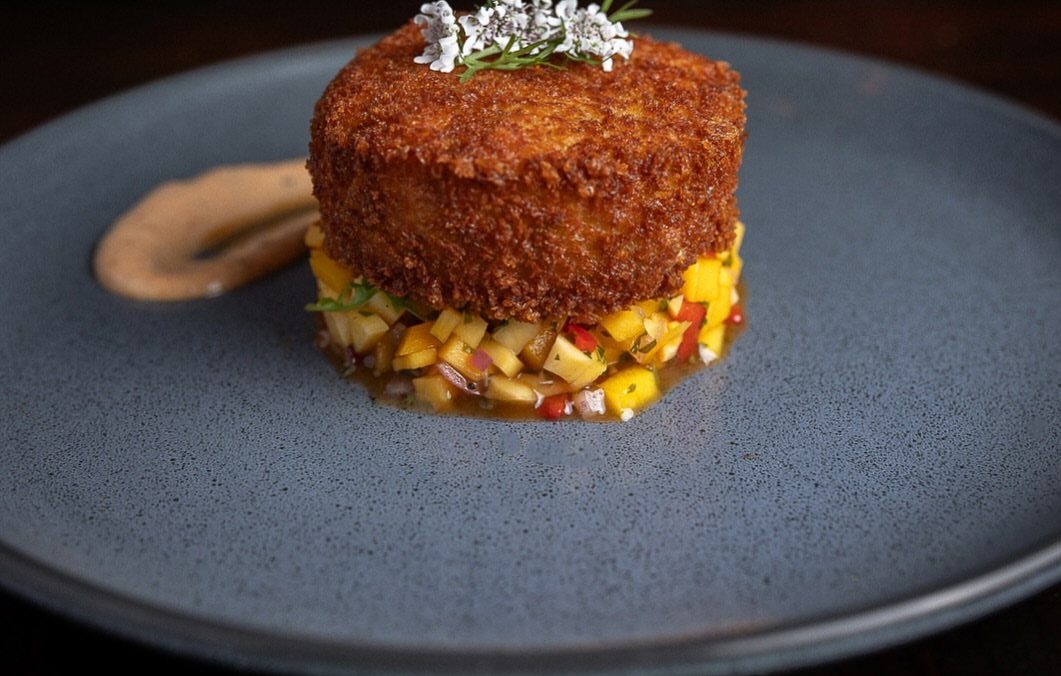 Golden-brown crab cake on mango salsa with a sauce swipe on grey plate