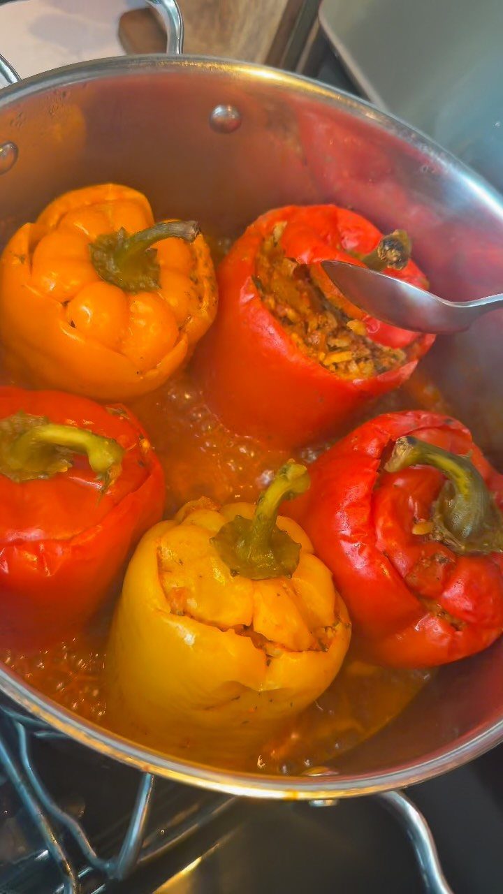 Stuffed red, orange, and yellow bell peppers simmering in a pot of sauce.