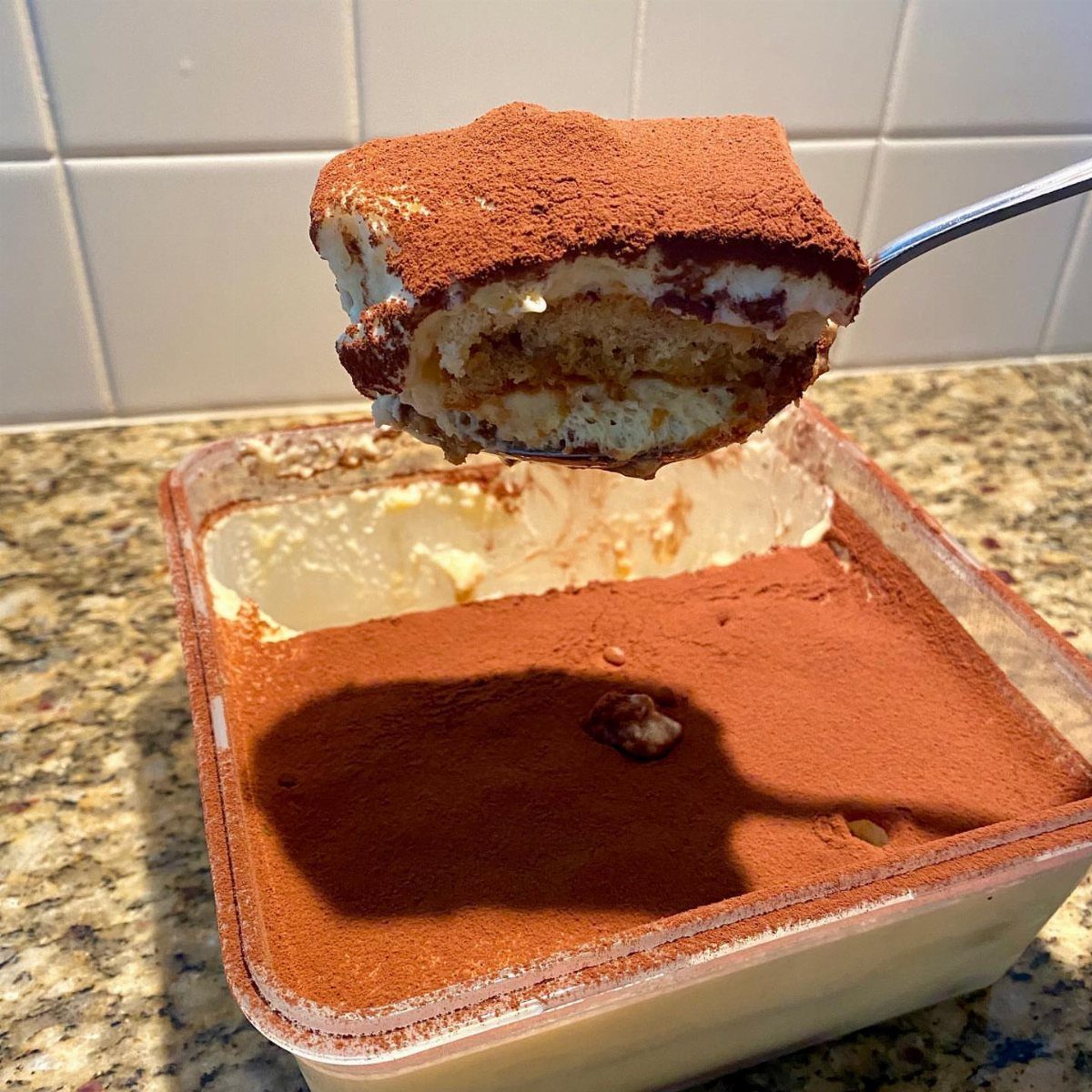 Tiramisu cake slice on a spoon held over a plastic container of dessert.