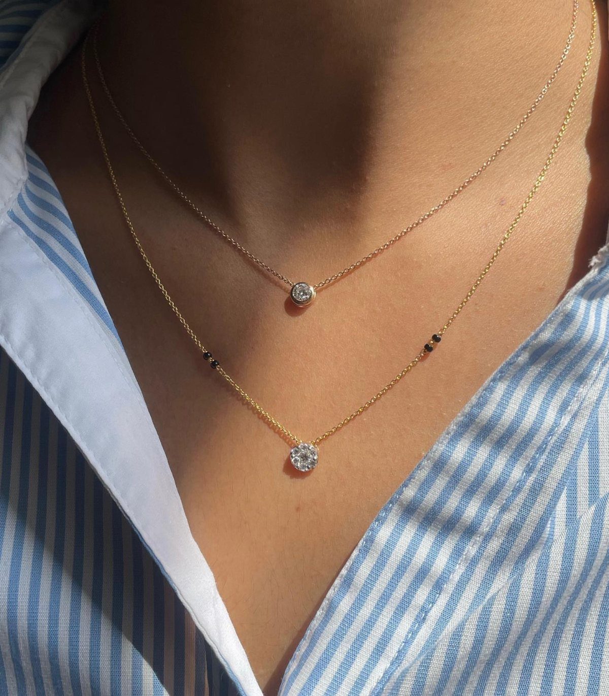 Two thin gold necklaces with small pendants worn over a striped blue shirt.