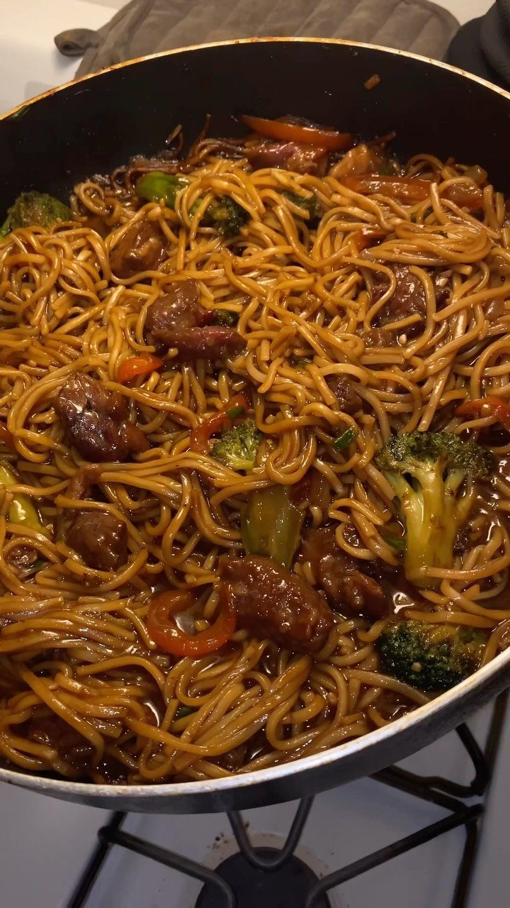 Beef lo mein with broccoli and red peppers in a black frying pan.