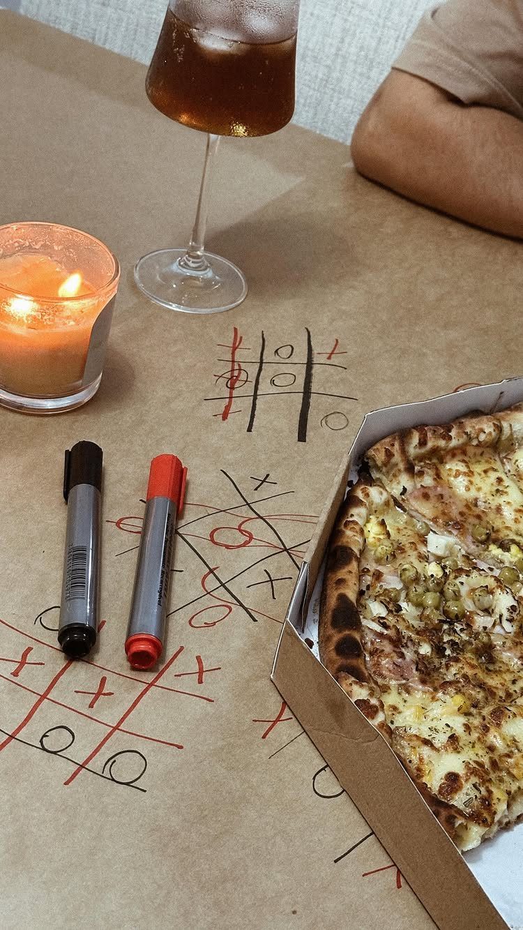 Tic-tac-toe games drawn on brown paper next to pizza, markers, and a candle.
