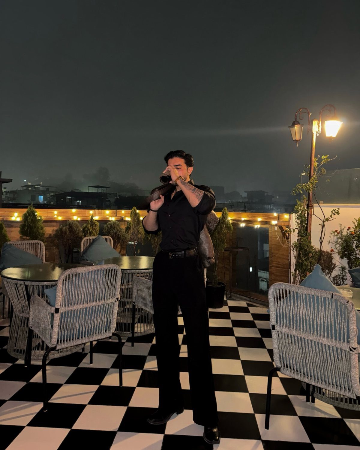 Man in black shirt and trousers posing on a checkered rooftop terrace at night.