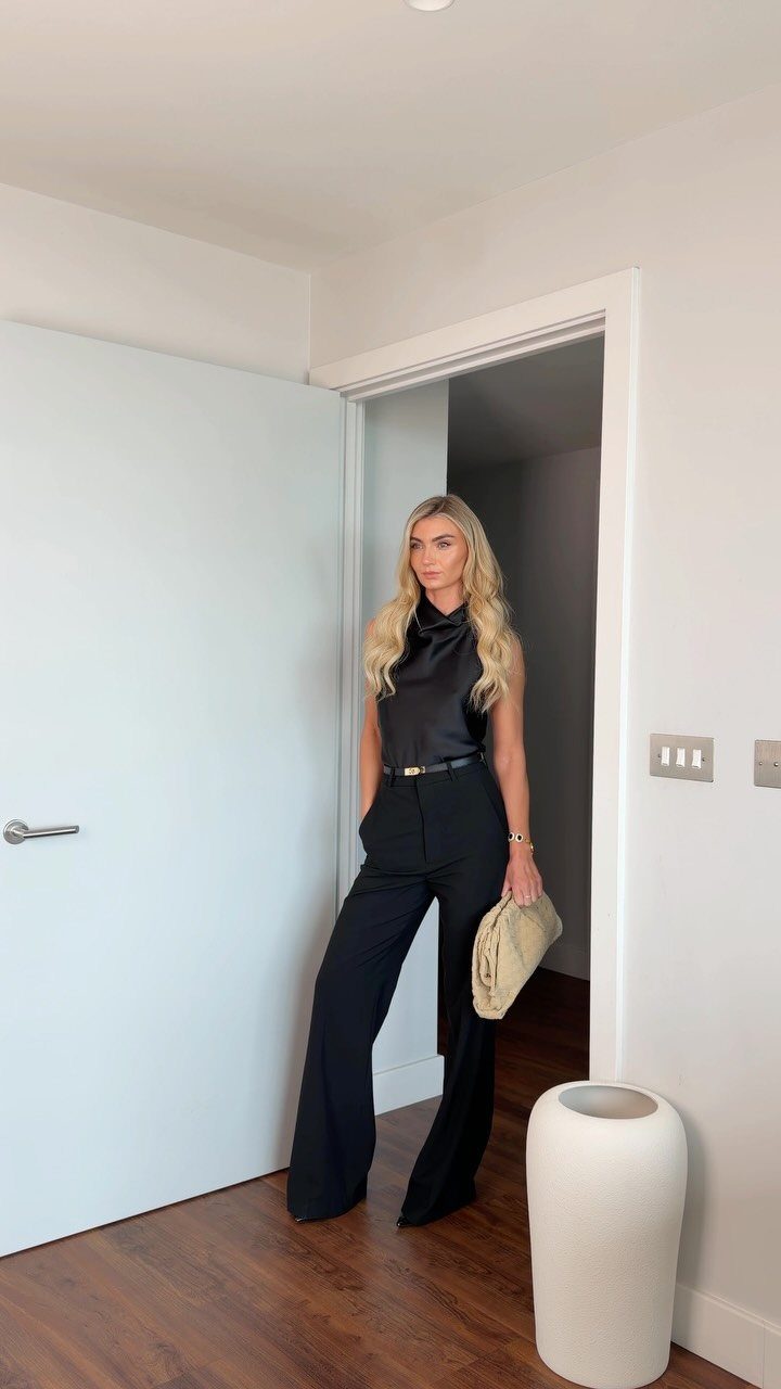 Woman wearing a black sleeveless top and wide-leg trousers with a beige clutch.