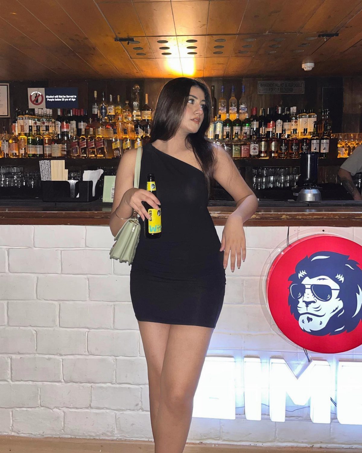 Woman in a black one-shoulder mini dress posing at a bar with drinks.