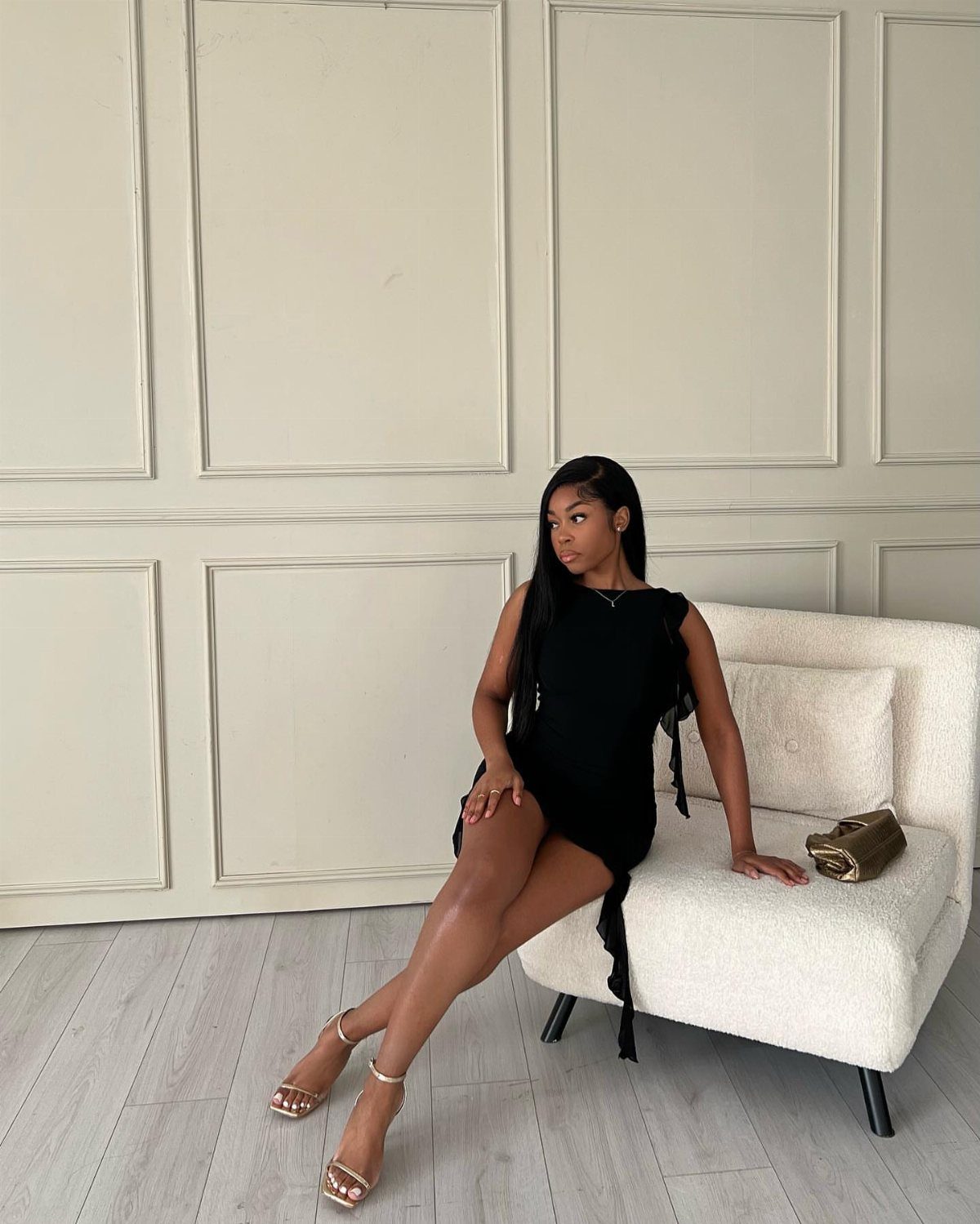 Woman in a short black dress and gold heels sitting on white chair