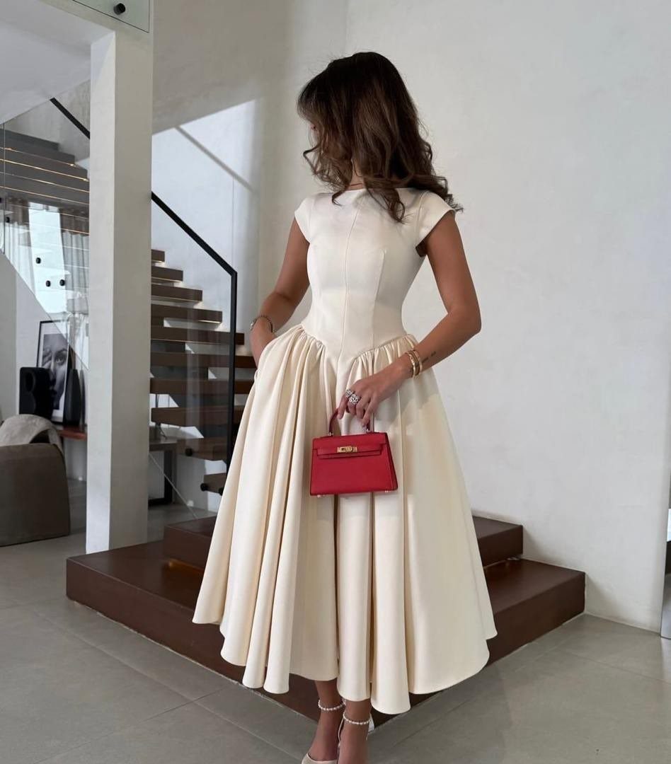 Woman in a cream midi dress holding a red handbag in a modern home.