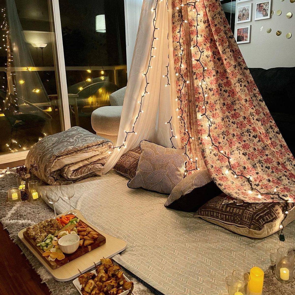Cozy indoor fabric fort with fairy lights, pillows, and a charcuterie board nearby.