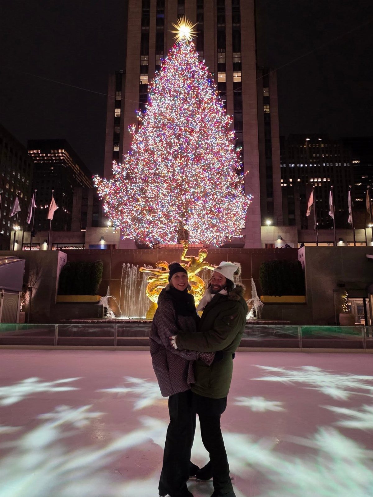 Couple hugging on an ice rink with a giant lit Christmas tree behind them.