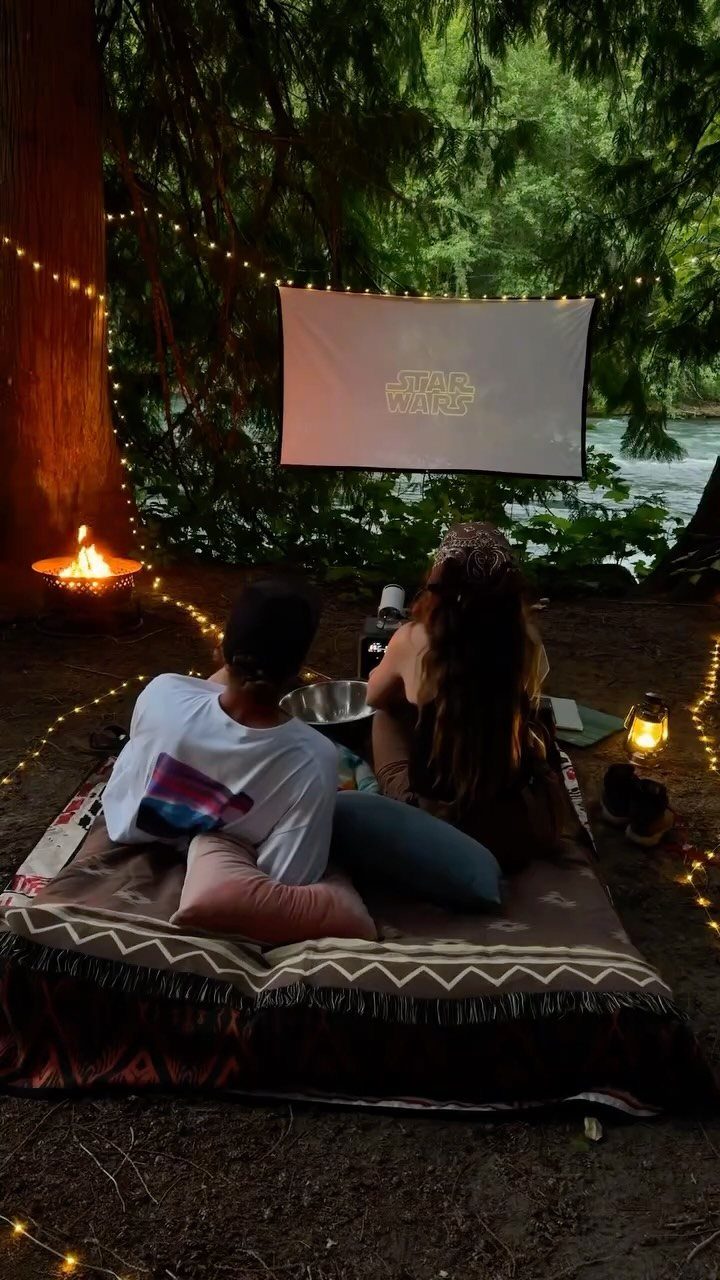 Couple watching a movie on a projector screen in a forest with string lights.