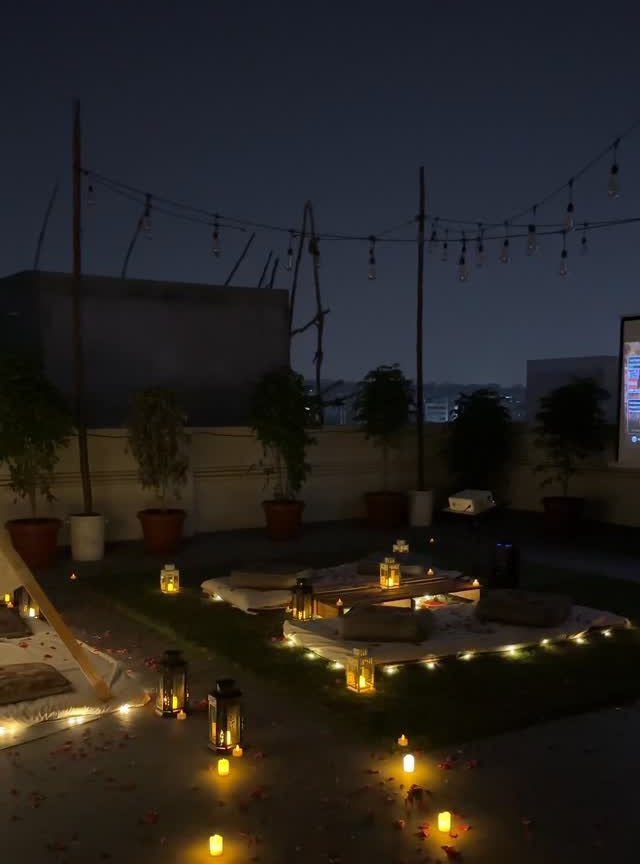 Romantic rooftop date setup with lanterns, cushions, and string lights at night.