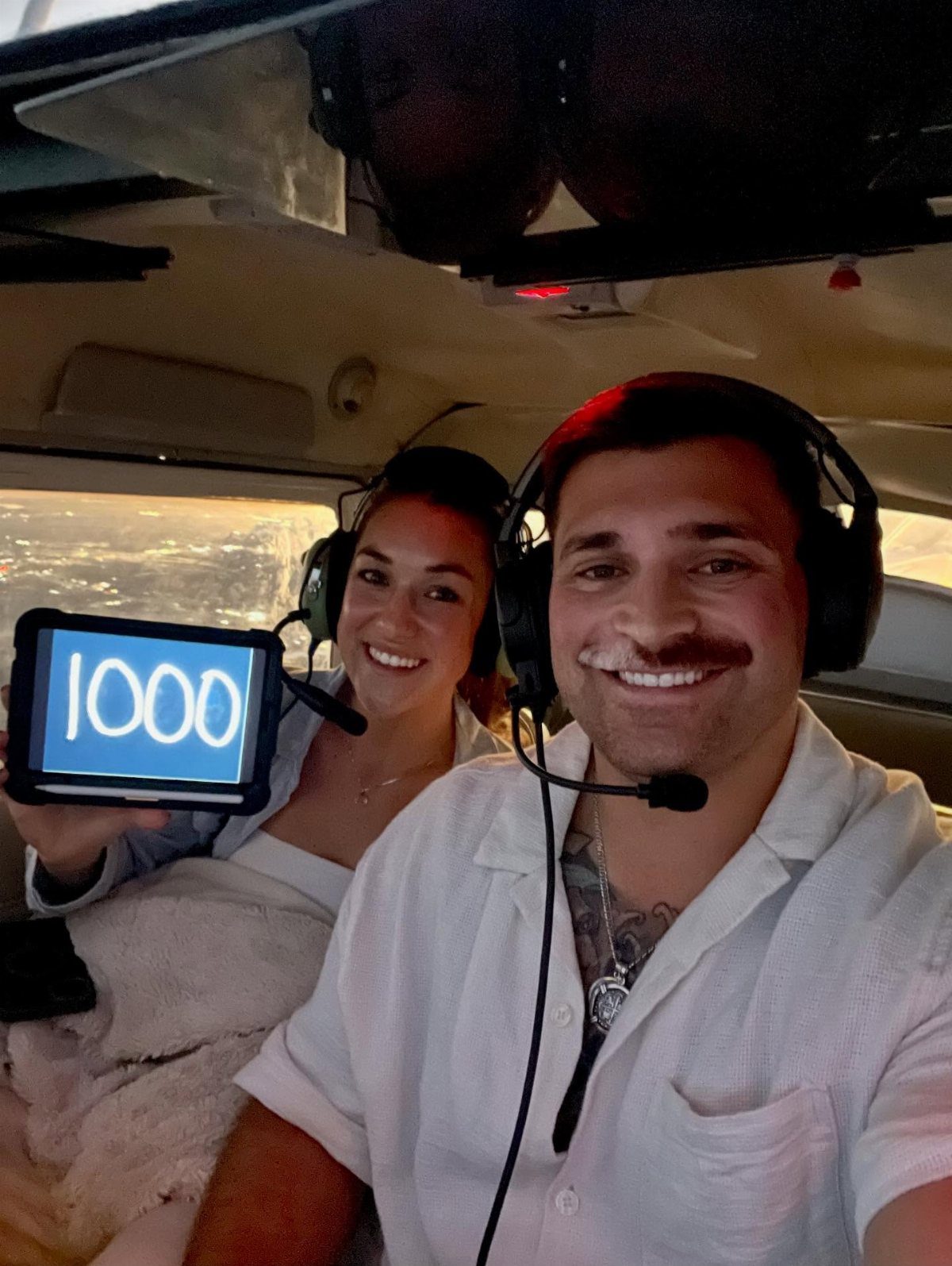 Smiling couple wearing aviation headsets inside a plane, holding a tablet showing 1000.
