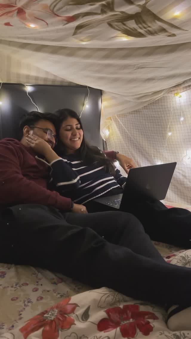 Couple cuddling under a blanket fort with string lights while using a laptop.
